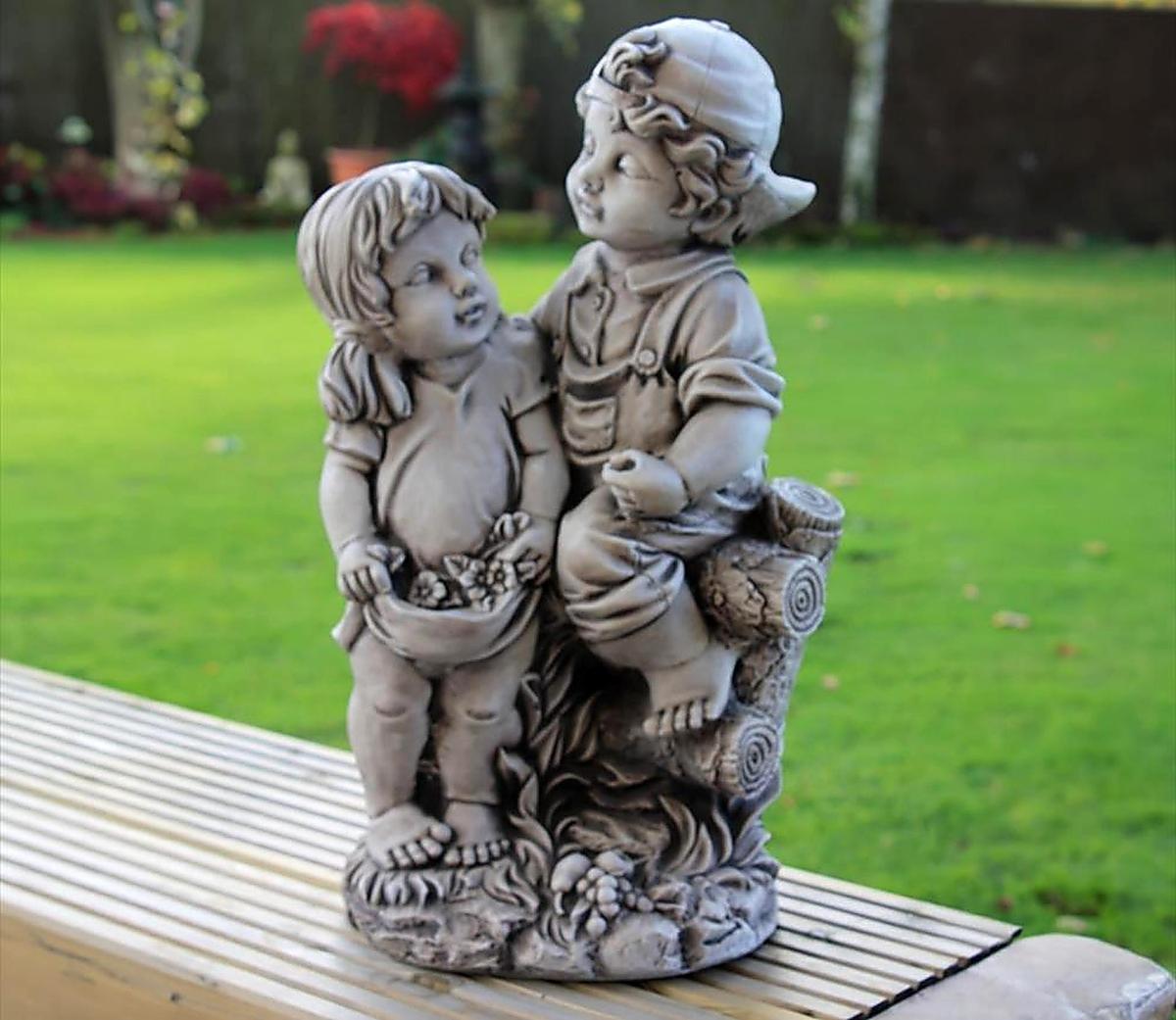 Stone garden statue depicting two children, one sitting on a log and the other holding a basket, set against a lush green lawn background. Ideal for outdoor decor.