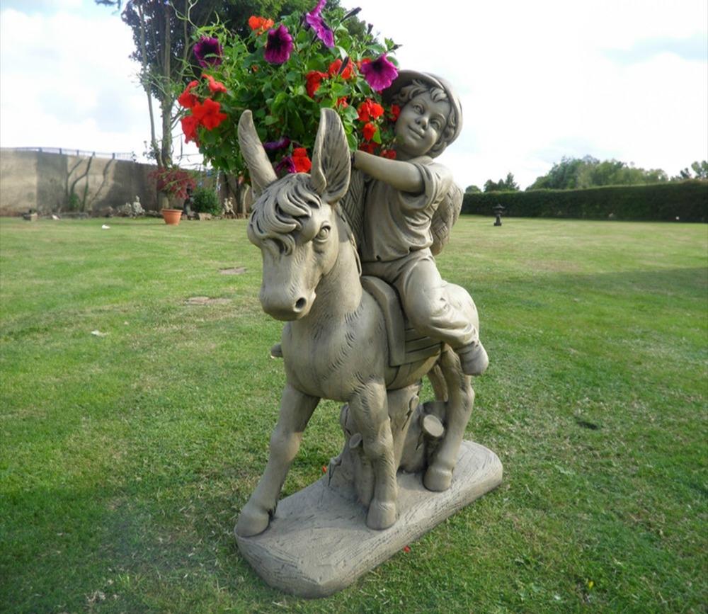 Stone garden ornament featuring a child on a donkey, carrying a vibrant planter with red and purple flowers. Ideal for adding charm to outdoor spaces.