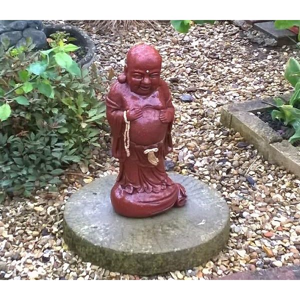 Red Laughing Buddha garden statue placed on a round stone pedestal, surrounded by gravel and greenery, symbolising happiness and prosperity in outdoor spaces.