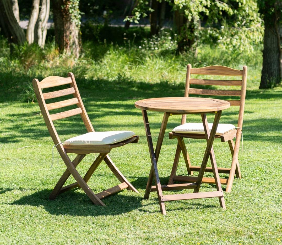 Wooden outdoor bistro set featuring two folding chairs with white cushions and a round table. Set on lush green grass with a backdrop of trees in summer.