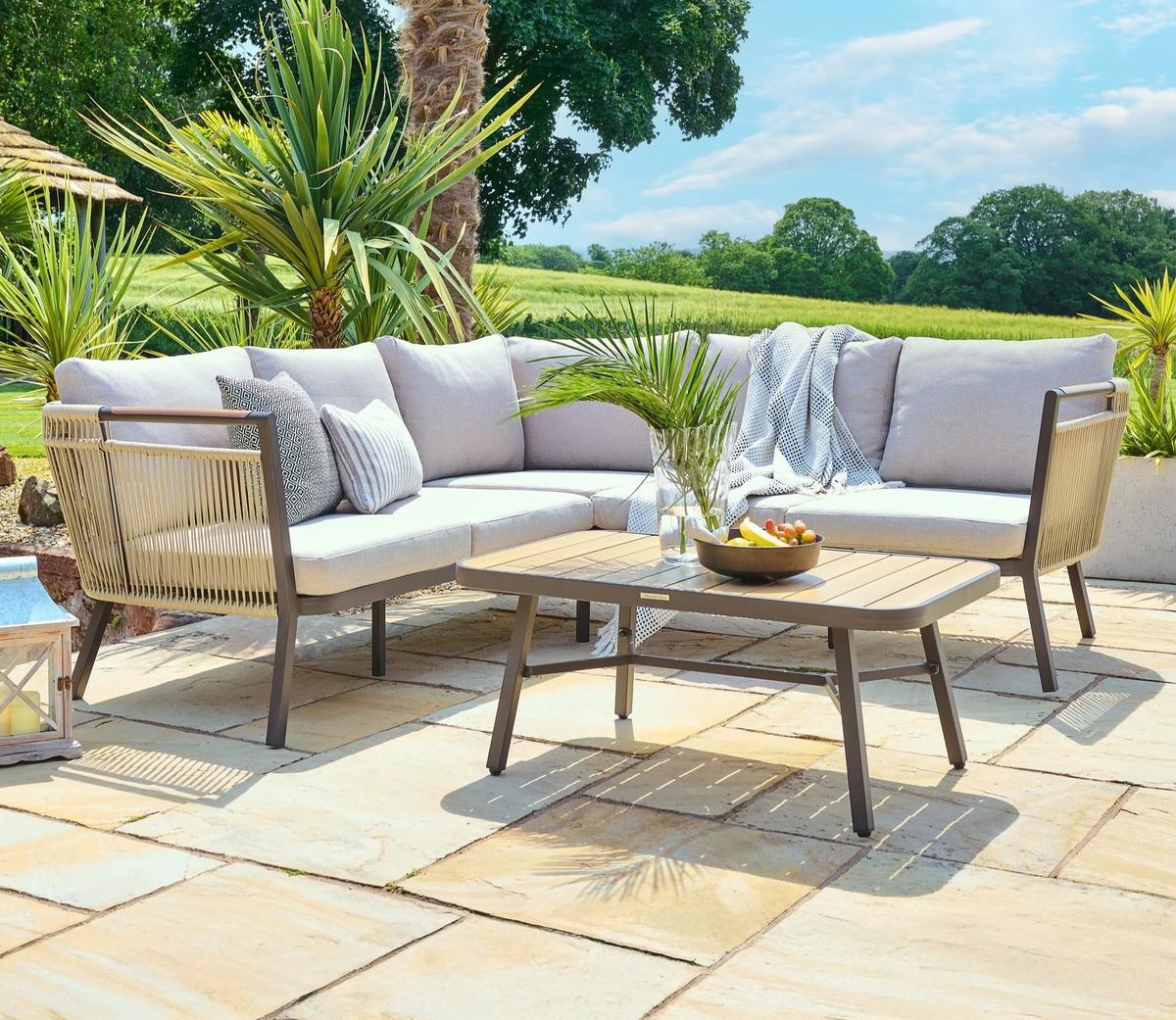 Outdoor sectional patio set with light grey cushions on a sunny stone terrace. Complemented by a wooden coffee table, lush green plants, and a scenic field view.