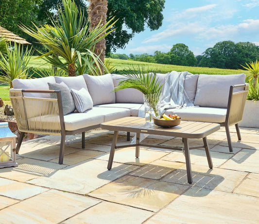 Outdoor sectional patio set with light grey cushions on a sunny stone terrace. Complemented by a wooden coffee table, lush green plants, and a scenic field view.