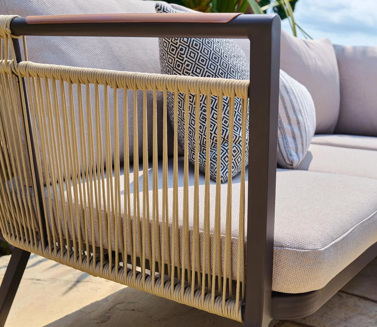 Outdoor beige sofa with decorative rope detailing on the armrest. Includes geometric-patterned cushions set against a sunny backdrop for a stylish patio setting.