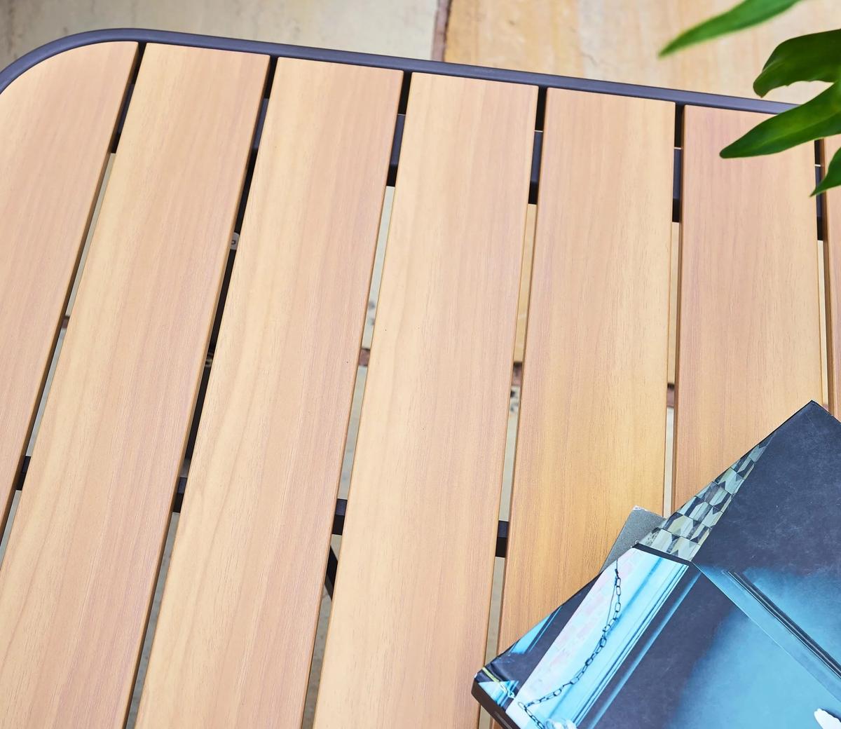 Wooden slatted table top with a smooth finish in a light colour, accompanied by an open magazine in the corner, and a small green plant on the side.