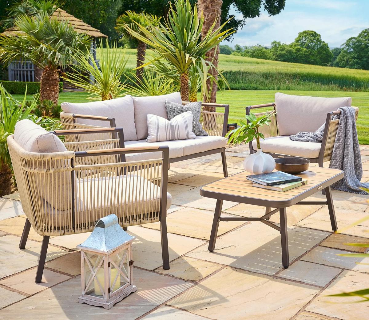 Outdoor sitting area featuring modern garden furniture set with beige cushions, stylish wooden coffee table, decorative lantern, and lush green surroundings on a sunny day.