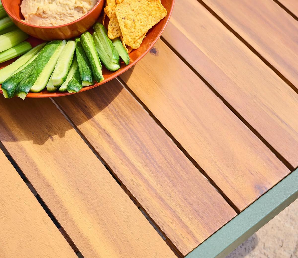 Close-up of a wooden outdoor table with a rich, natural finish, showcasing a vibrant orange bowl. The bowl contains sliced cucumbers, tortilla chips, and hummus.