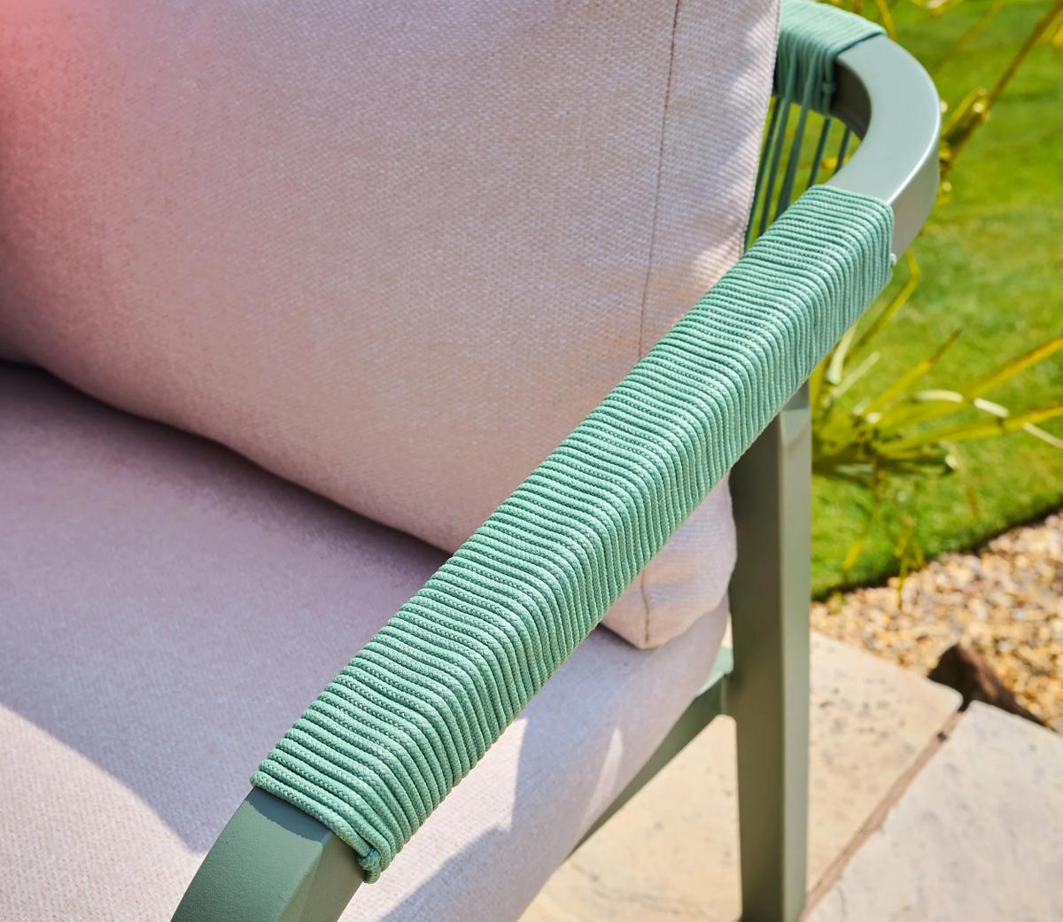 Close-up of a garden chair with a green textured armrest and soft pink cushion, showcasing modern outdoor furniture design against a vibrant green lawn and gravel path.