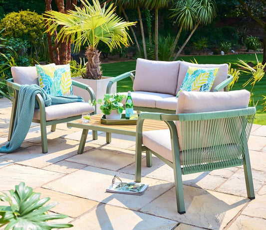 Modern outdoor patio furniture set with two armchairs, a sofa, and a coffee table on a sunny stone patio. Features green cushions and vibrant tropical cushions.