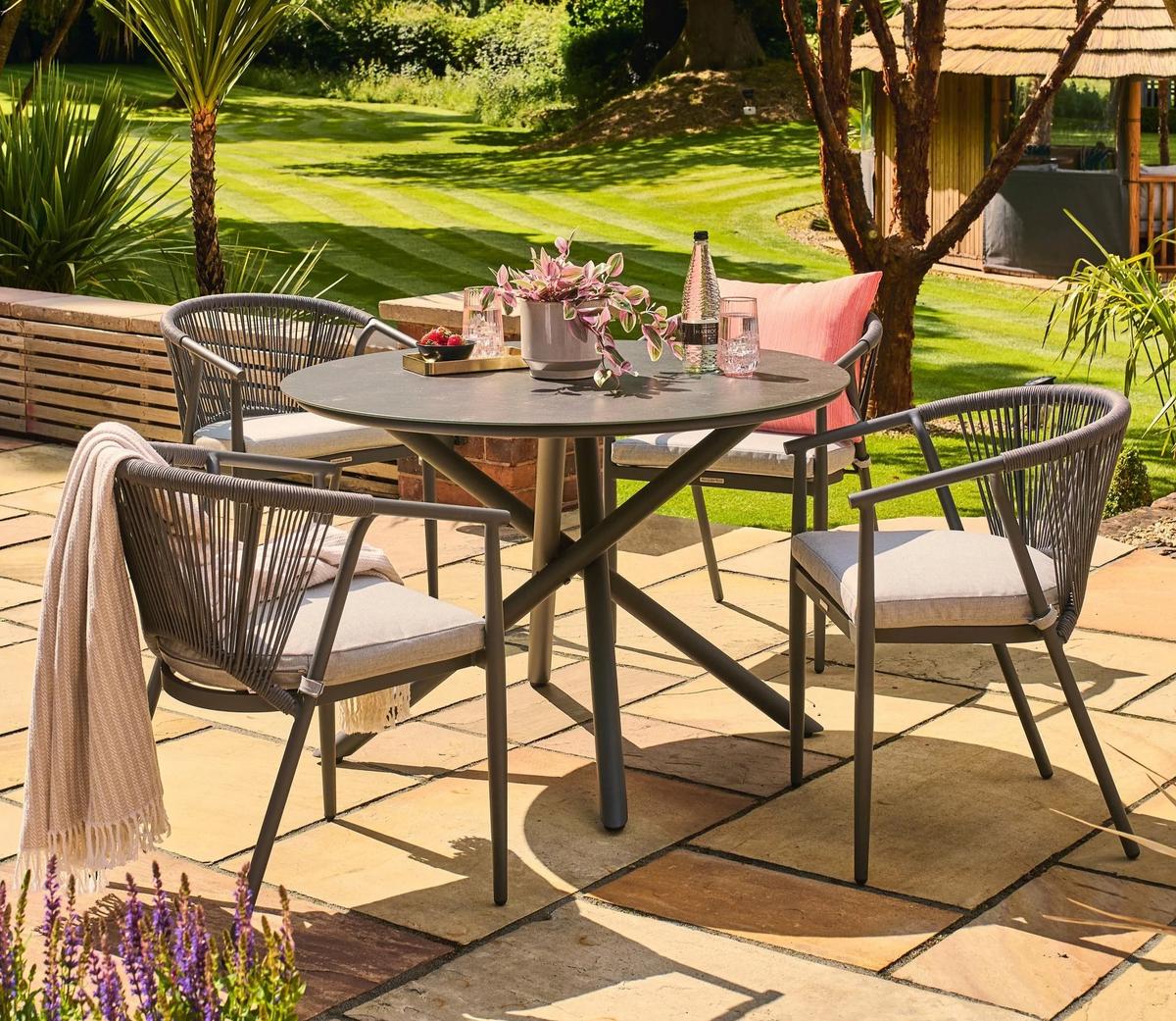 Four-piece grey outdoor dining set on a sunny patio, featuring a round table with a decorative plant, surrounded by armchairs, and a scenic garden backdrop.
