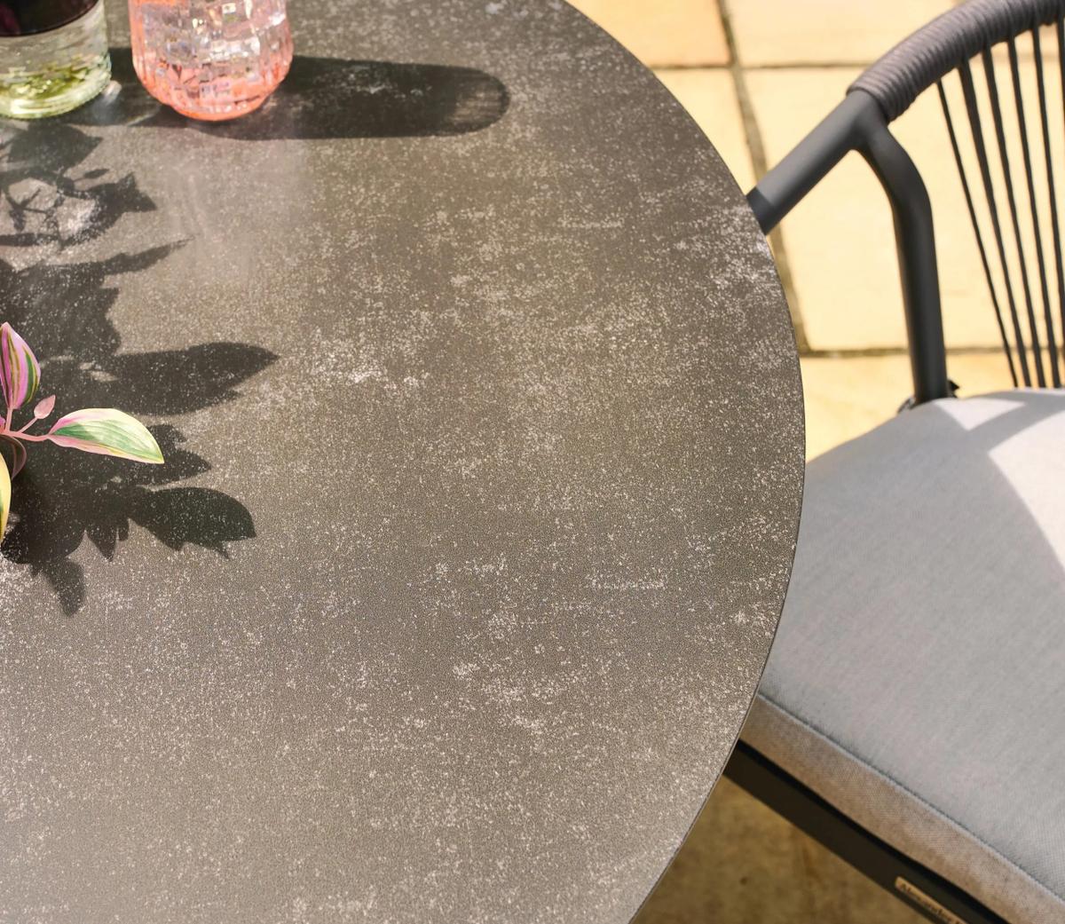 A close-up image of a round outdoor patio table with a textured, dark surface, paired with a grey cushion chair and decorative plants, set against a tiled floor backdrop.