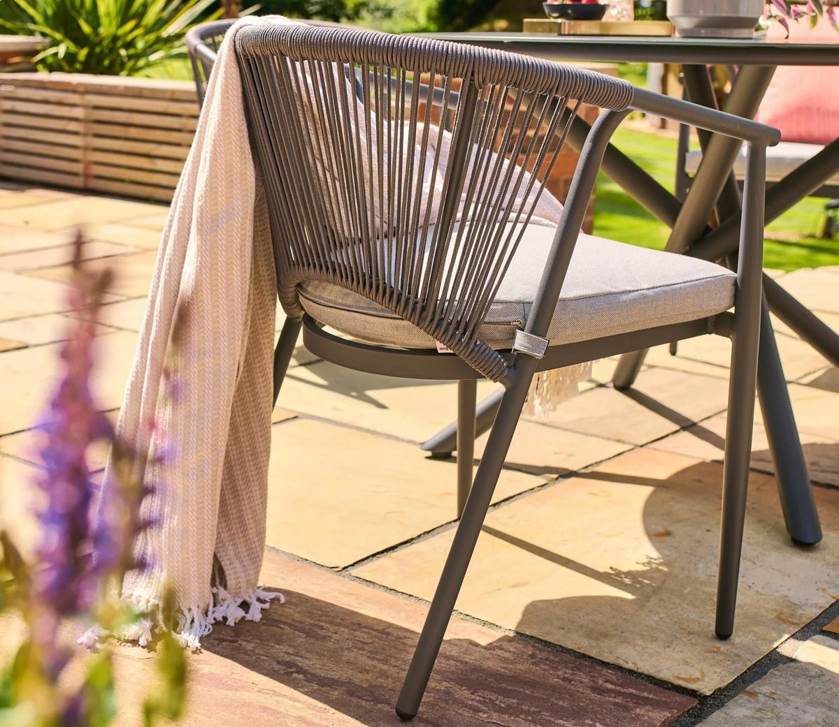 Stylish outdoor dining chair with a taupe cushion and rope design, adorned with a beige throw, on a sunny patio, beside a round metal table.