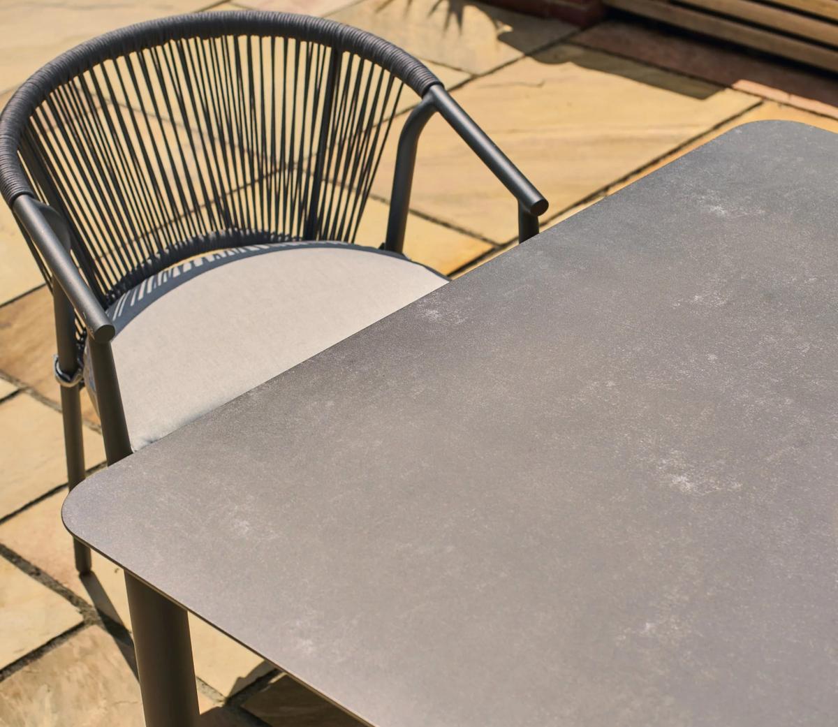 A modern patio setting featuring a dark, textured outdoor table with a rounded corner. Accompanied by a grey chair with a rope backrest on a stone-tiled terrace.
