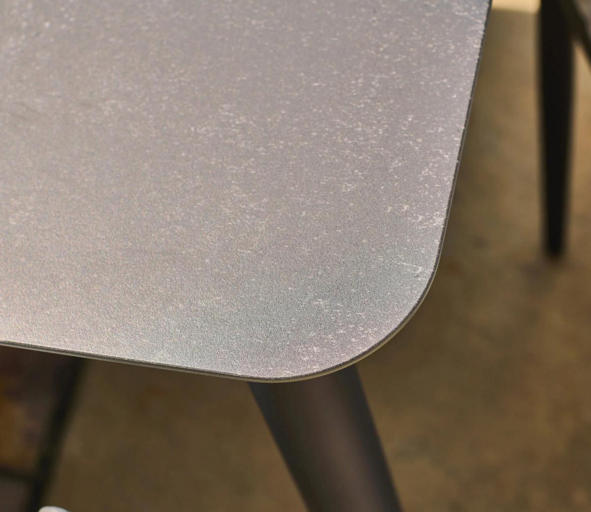 Close-up of a rectangular metal tabletop corner with a smooth matte finish, showing the subtle texture and rounded edge. The table stands on black angled legs.