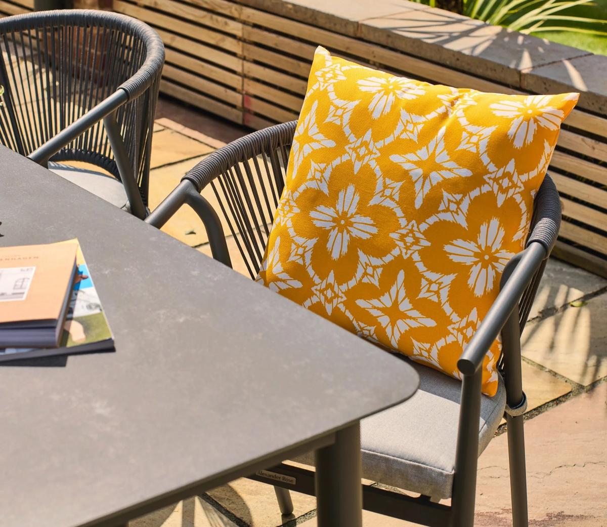Outdoor patio scene showing a stylish yellow cushion with white floral patterns on a grey chair beside a modern black metal dining table for garden settings.