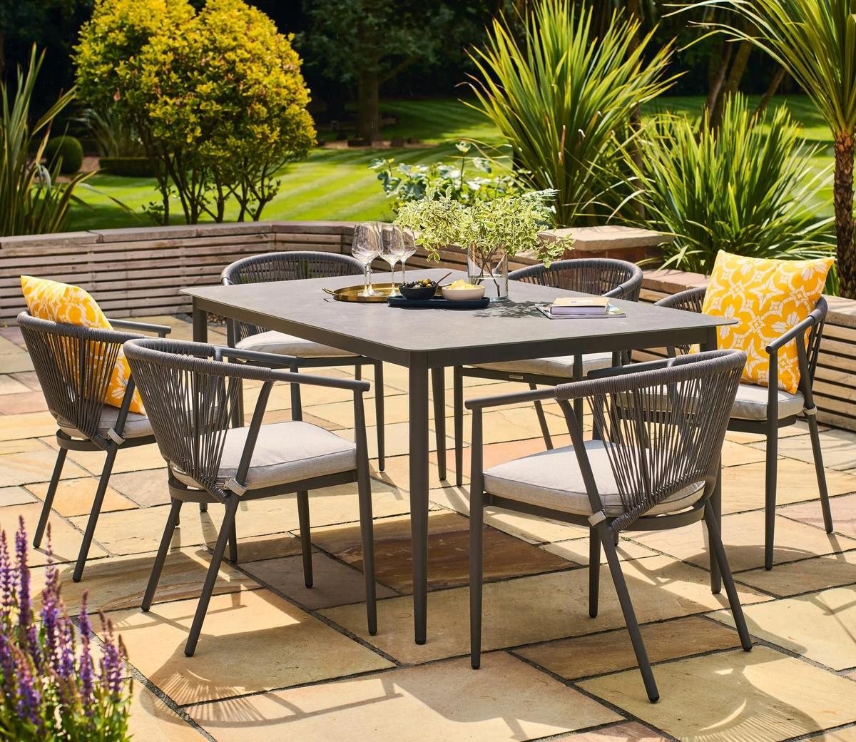 Outdoor patio set featuring six grey wicker chairs with cushions and a rectangular table, set on a stone patio with greenery and yellow accents on pillows.