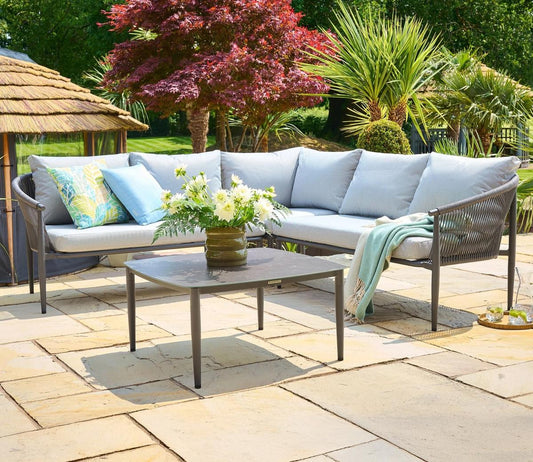 Modern outdoor seating set featuring a grey corner sofa with light blue cushions, a stylish coffee table, decorative pillows, and lush greenery in the background.