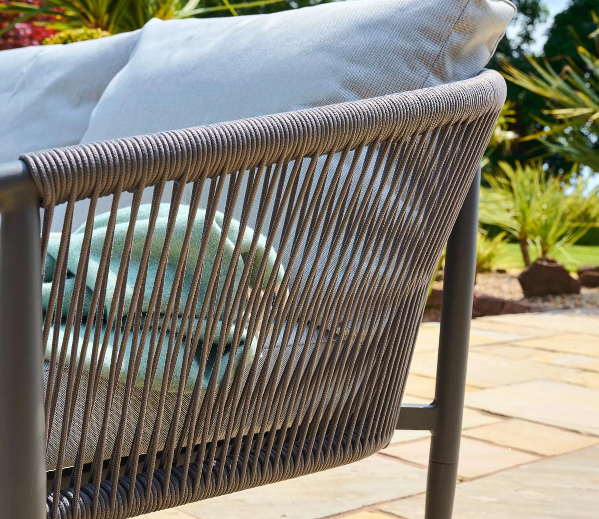 Outdoor rope-woven sofa detail showcasing its durable grey frame and plush light blue cushions, set against a garden landscape with vibrant greenery.
