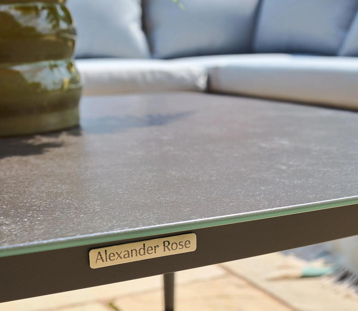 Close-up of a garden table with a metallic surface and the Alexander Rose branding, accompanied by a green vase, set against a blurred outdoor sofa background.