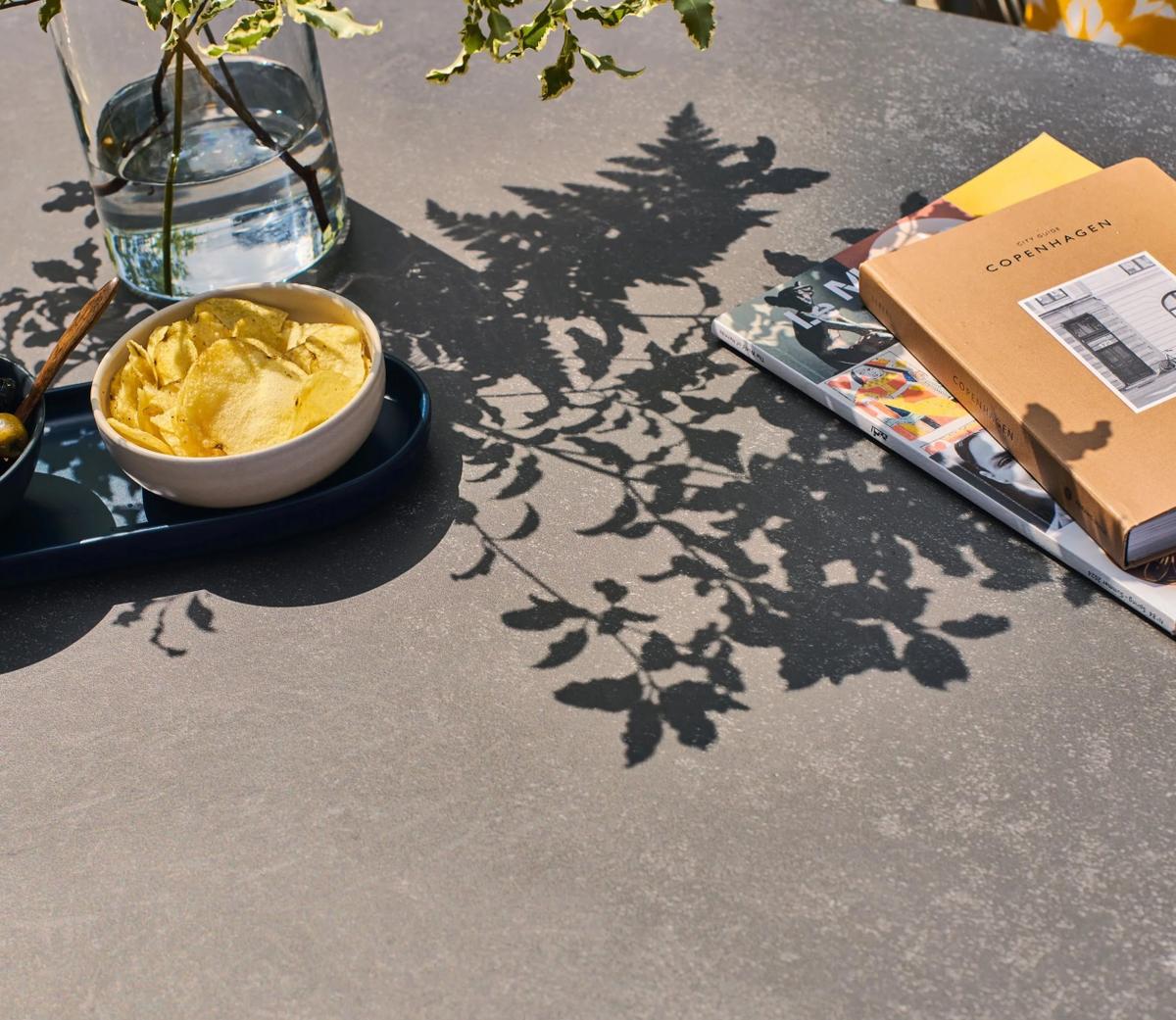 A modern coffee table with a sleek dark surface is adorned with a glass vase of water, delicate leaf shadows, a bowl of crisps, and fashion magazines on Copenhagen.
