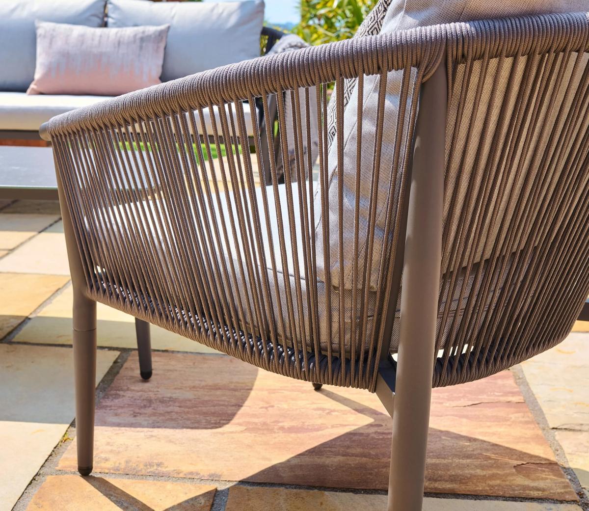 Outdoor armchair with a woven rattan design and beige cushions on a sunny patio. The chair features a modern, elegant style suitable for garden settings.