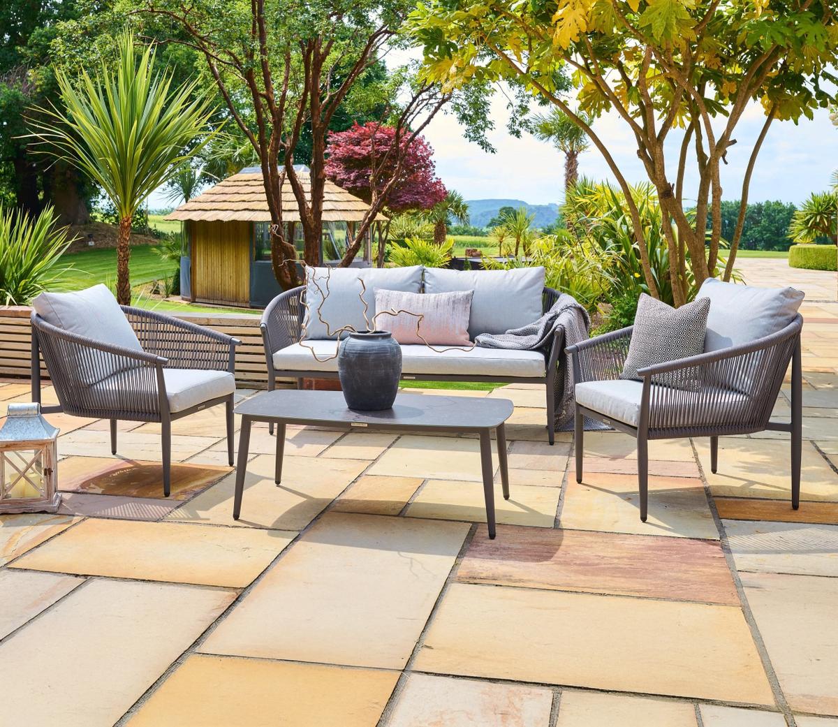 Outdoor garden seating set on a stone patio with two grey armchairs, a sofa, and a coffee table. Surrounded by lush greenery and trees, creating a serene environment.
