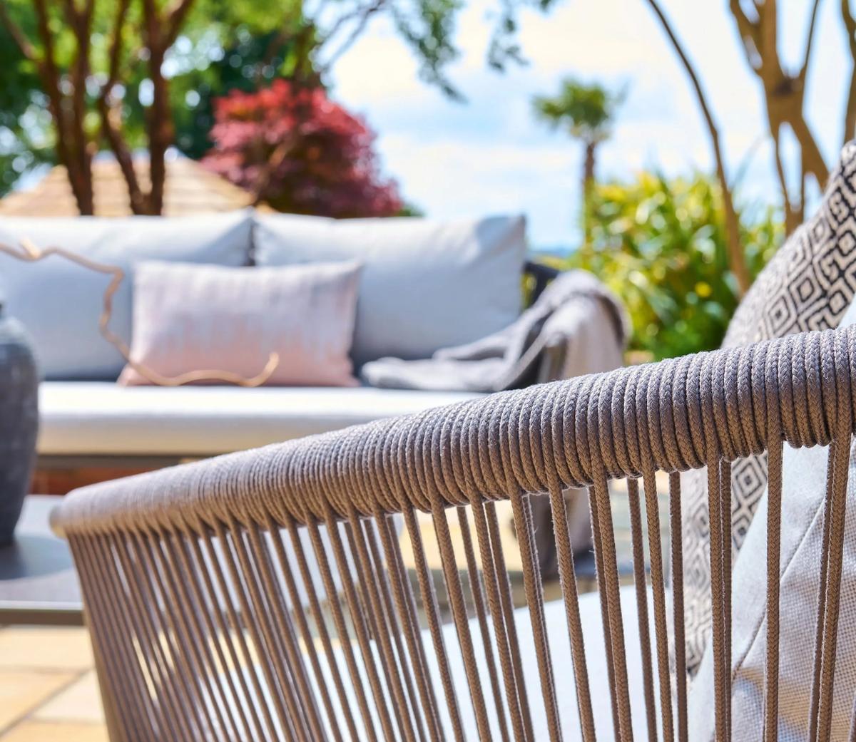 Close-up of the intricate rope weave detail on a stylish outdoor chair, set in a garden with lush greenery and a cushioned sofa in the background. Perfect for modern patios.