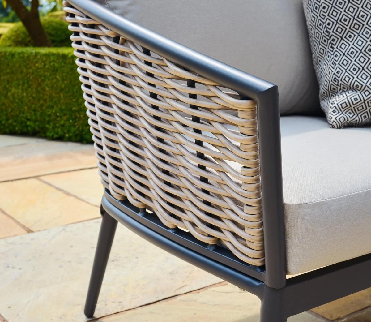 Close-up view of a modern outdoor chair with grey metal frame and beige rattan weave. Soft beige cushion and black-patterned pillow are visible against a patio backdrop.