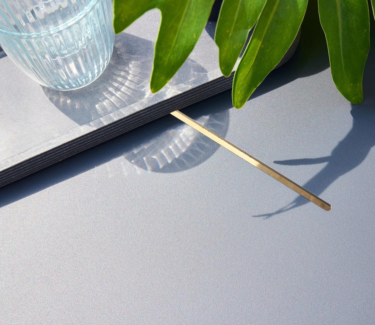A close-up of a textured grey rectangular tile with a gold metallic accent resting on it. A clear ribbed glass and green plant leaves create delicate, intricate shadows.