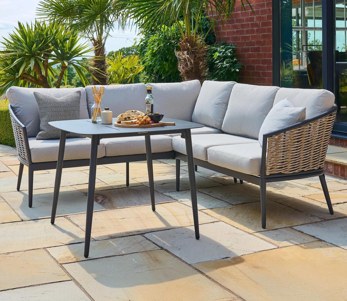 Outdoor corner sofa set with grey cushions and stylish woven sides, arranged on a sunlit patio surrounded by lush green plants, featuring a dining table set.