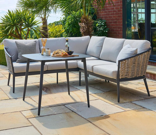 Outdoor corner sofa set with grey cushions and stylish woven sides, arranged on a sunlit patio surrounded by lush green plants, featuring a dining table set.