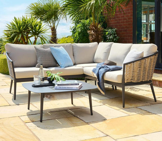Outdoor patio with stylish beige rattan sofa set, featuring plump cushions and a sleek coffee table on a sunny terrace. Surrounded by lush green palm trees.