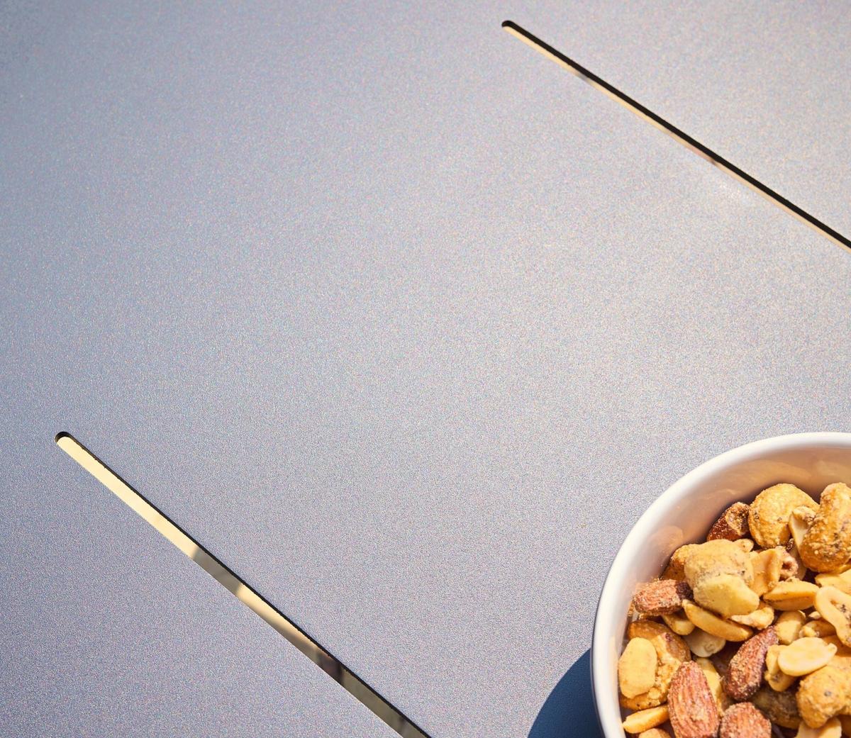 A close-up of a white bowl filled with assorted nuts and snacks, placed on a sleek grey table with narrow gaps, under natural sunlight.
