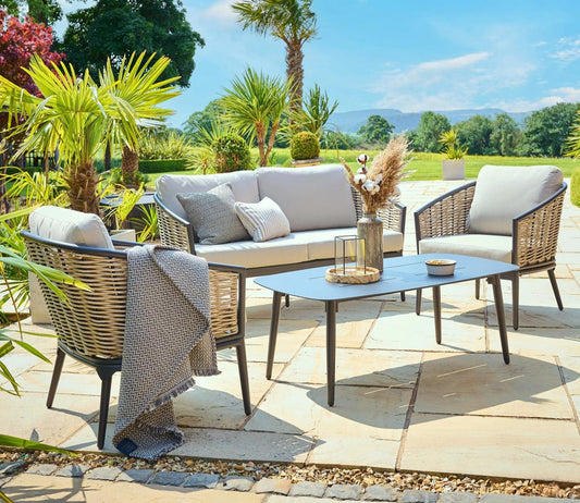 Outdoor patio set featuring a sofa, two armchairs, and a coffee table on stone tiles. Surrounded by lush greenery and gardens under a clear blue sky.