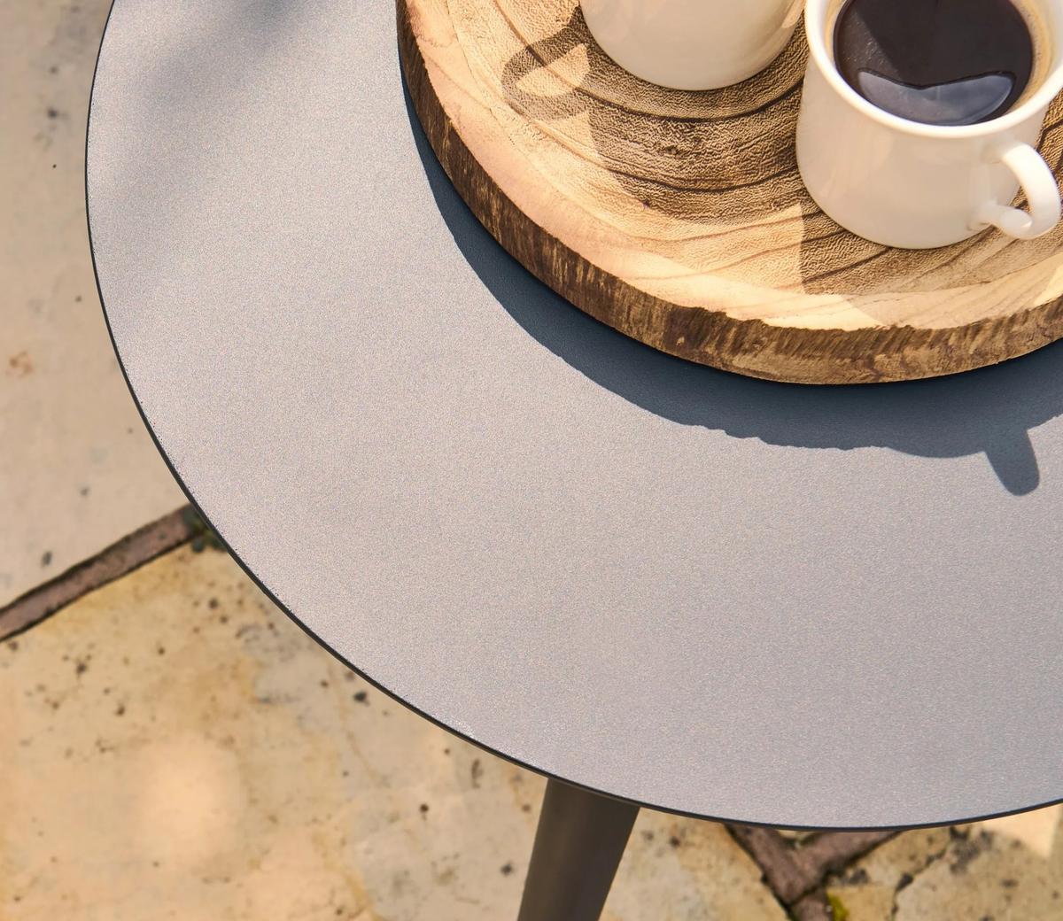 A minimalist outdoor table with a smooth grey surface, featuring a round wooden tray holding two cups of coffee, set on a patio with natural light.
