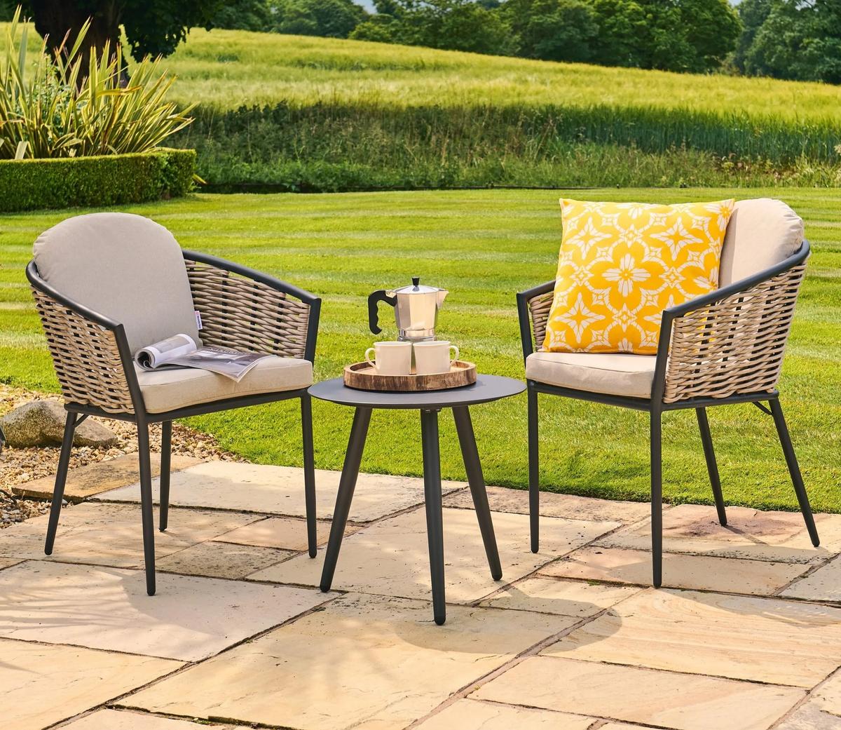 Outdoor patio set featuring two rattan chairs with beige cushions and a stylish round table. Set on a stone patio overlooking a lush, well-maintained garden.