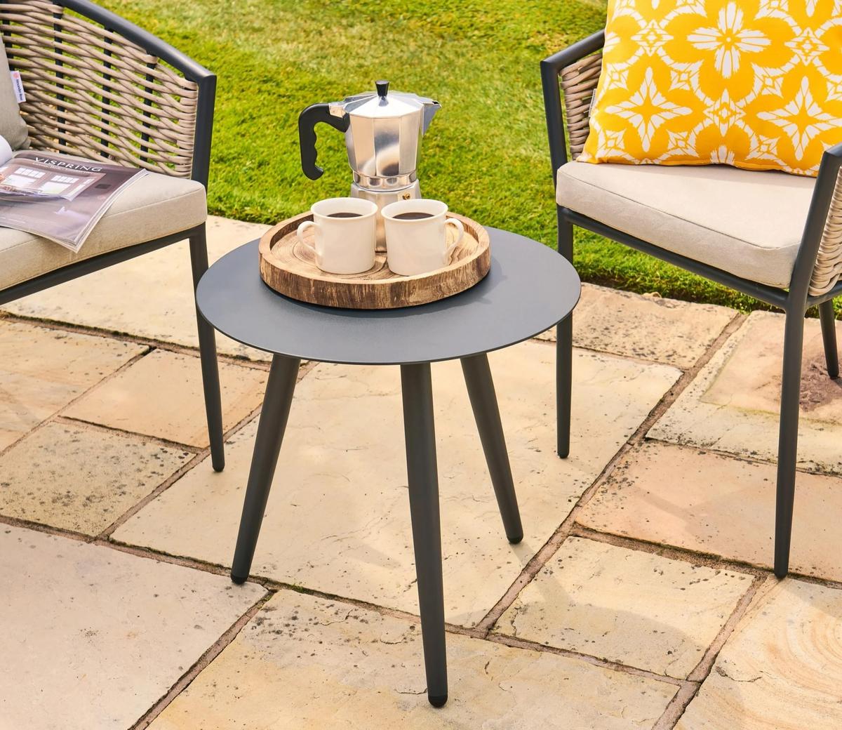 A modern patio set featuring a round grey metal table with a moka pot and two cups on a wooden tray, flanked by cushioned wicker chairs with a yellow pillow.