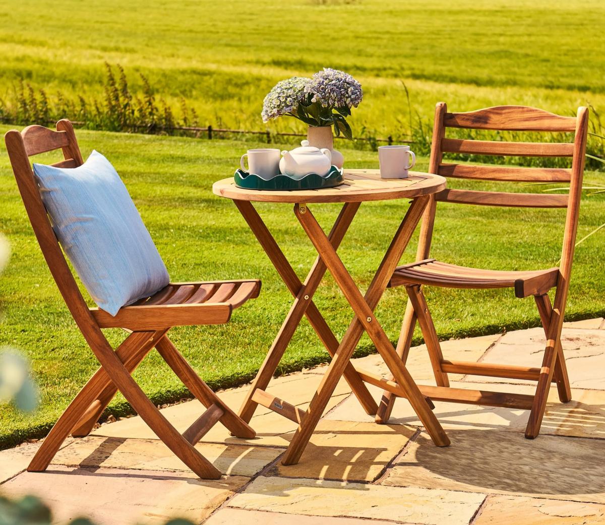 Wooden bistro set with two foldable chairs and round table, adorned with a teapot and flowers. Ideal for garden patios, offering a cosy outdoor seating area.