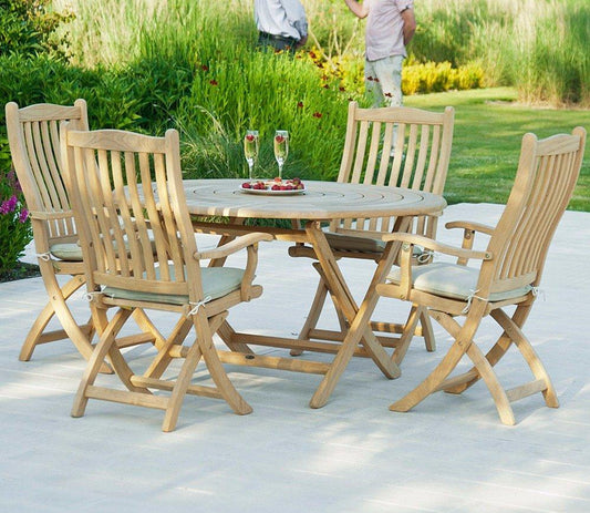 A wooden outdoor dining set on a patio featuring a round table and five cushioned chairs. The setting is enhanced by a lush green garden backdrop.