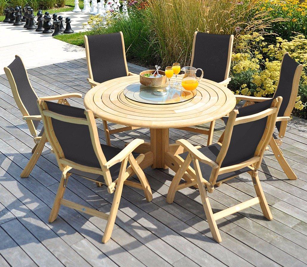 Wooden outdoor dining set with a round table and six reclinable chairs on a patio, surrounded by lush garden plants, featuring drinks and a salad bowl.