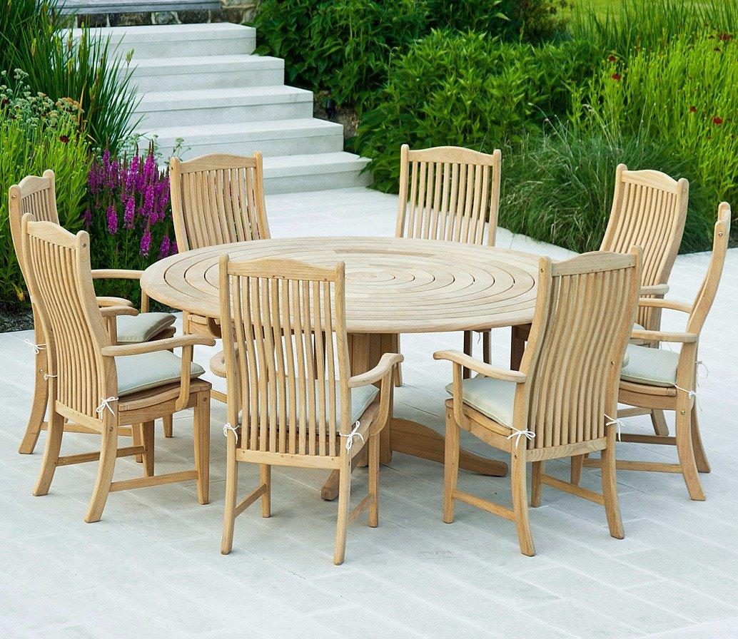 Round teak outdoor dining set with eight cushioned chairs on a patio. Surrounded by vibrant green plants and purple flowers, offering a serene garden setting.