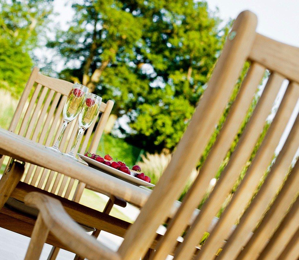 Wooden patio dining set with tall chairs and a table featuring two glasses of champagne and a plate of fresh raspberries, set in a sunny outdoor garden setting.