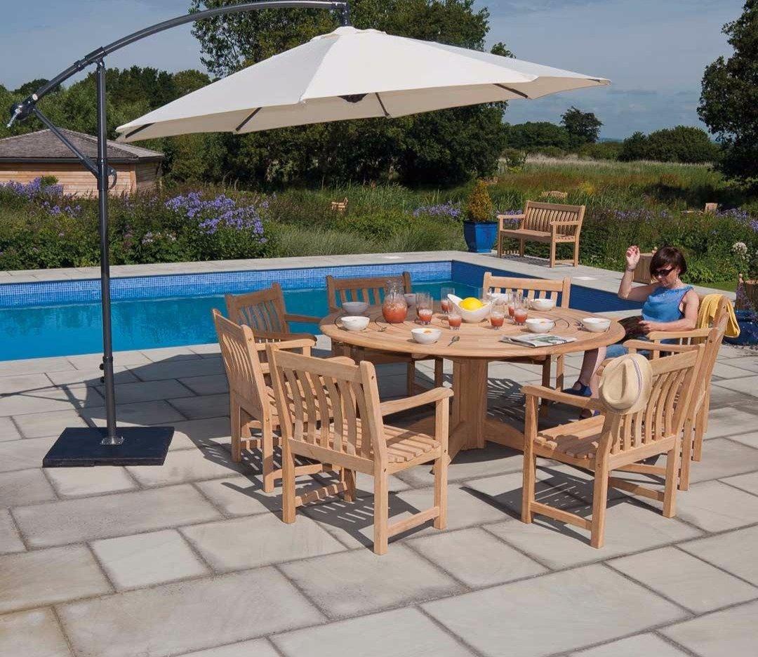 Outdoor wooden dining set with six chairs and a round table, shaded by a large white parasol beside a pool. Ideal for summer gatherings in a garden setting.