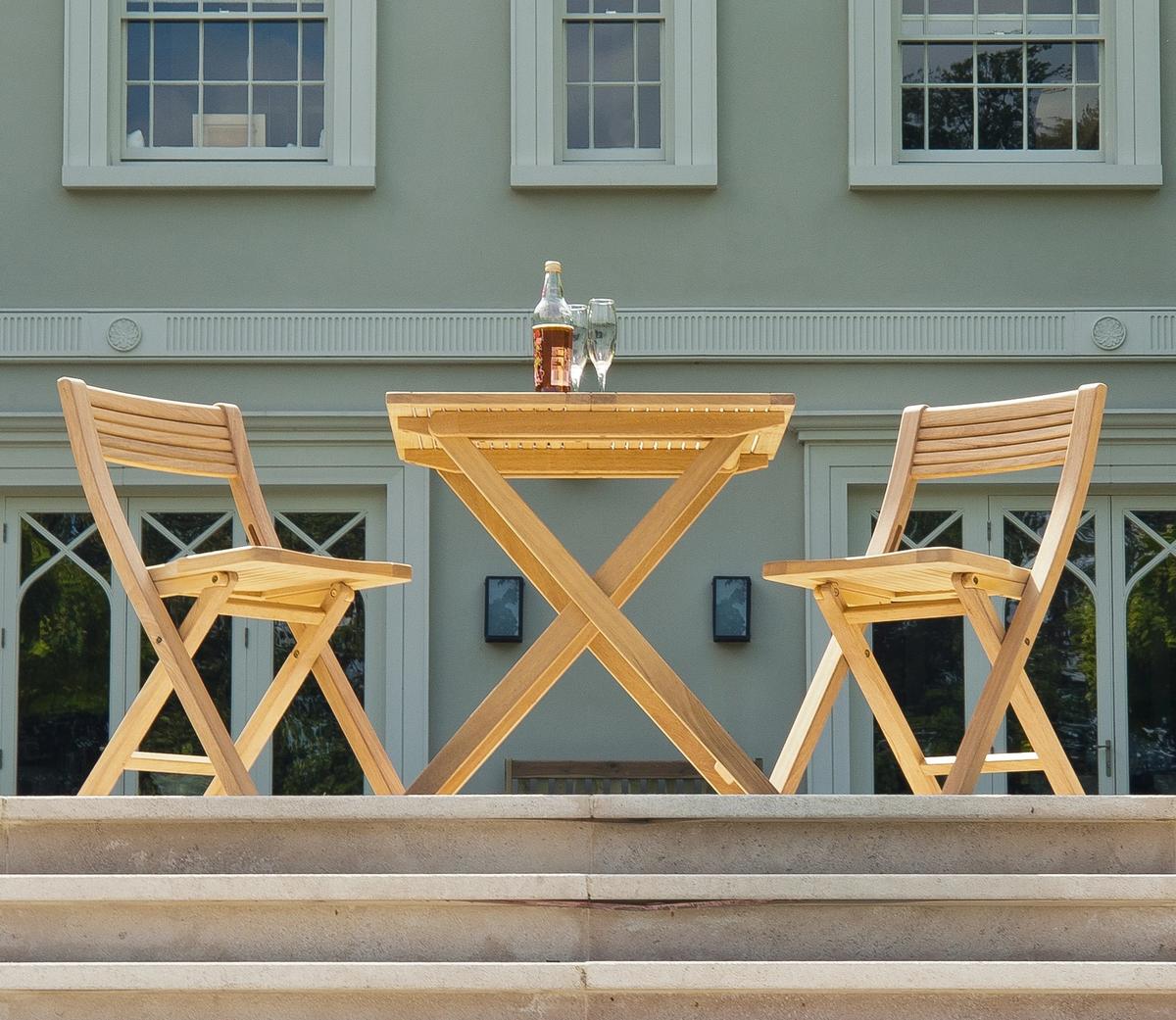 Two wooden outdoor chairs and a matching table on a stone patio, set with a drink and glasses, offering a stylish setting for garden relaxation in front of a classic house.