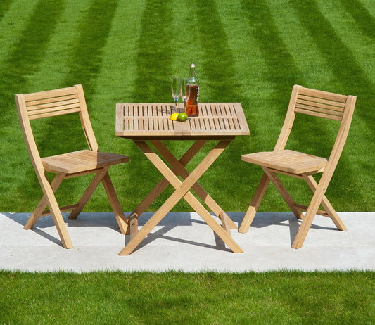 A wooden patio set with a slatted table and two chairs on a stone path, surrounded by vibrant green grass. Includes two glasses, a soda bottle, and citrus fruits.