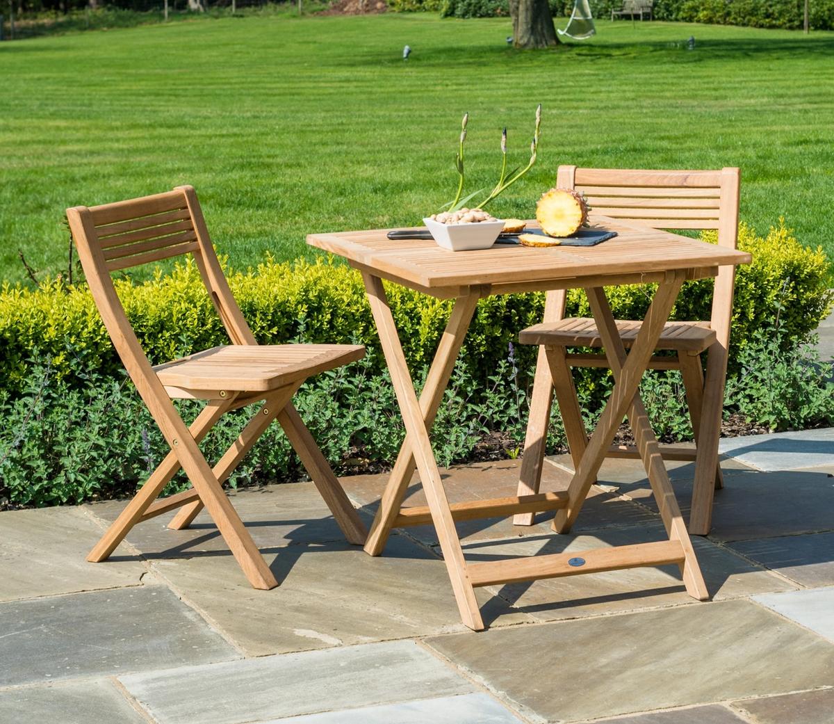 A natural teak bistro set featuring two foldable chairs and a square table, perfect for outdoor dining. The set is displayed on a stone patio with lush green grass.