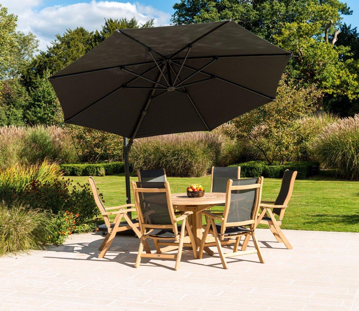 Outdoor patio set with six wooden chairs and a round wooden table, adorned with a large, dark umbrella, placed on a stone patio in a lush garden setting.