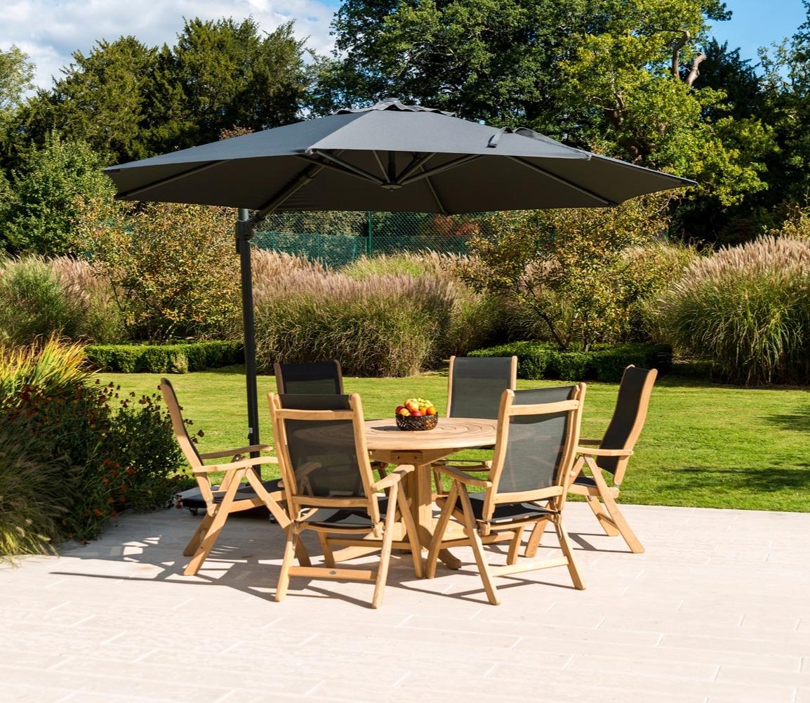 Wooden outdoor dining set with a round table and six chairs under a large grey parasol on a stone patio, surrounded by lush green garden foliage and trees.