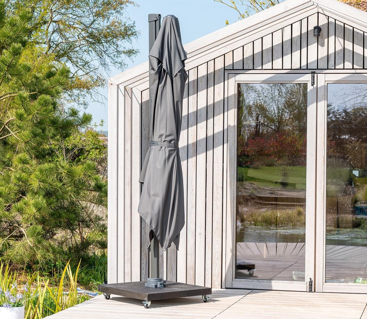 Closed grey cantilever parasol on wheeled base beside modern garden building, compact and practical garden shade solution for patios and outdoor spaces.