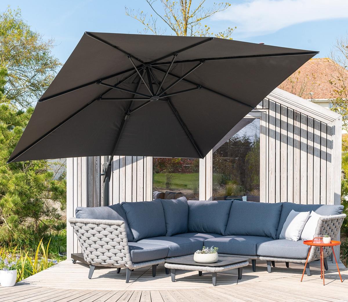 Large cantilever parasol fully open above outdoor corner sofa, creating shaded seating area on a wooden deck beside a modern garden room and landscaped garden.