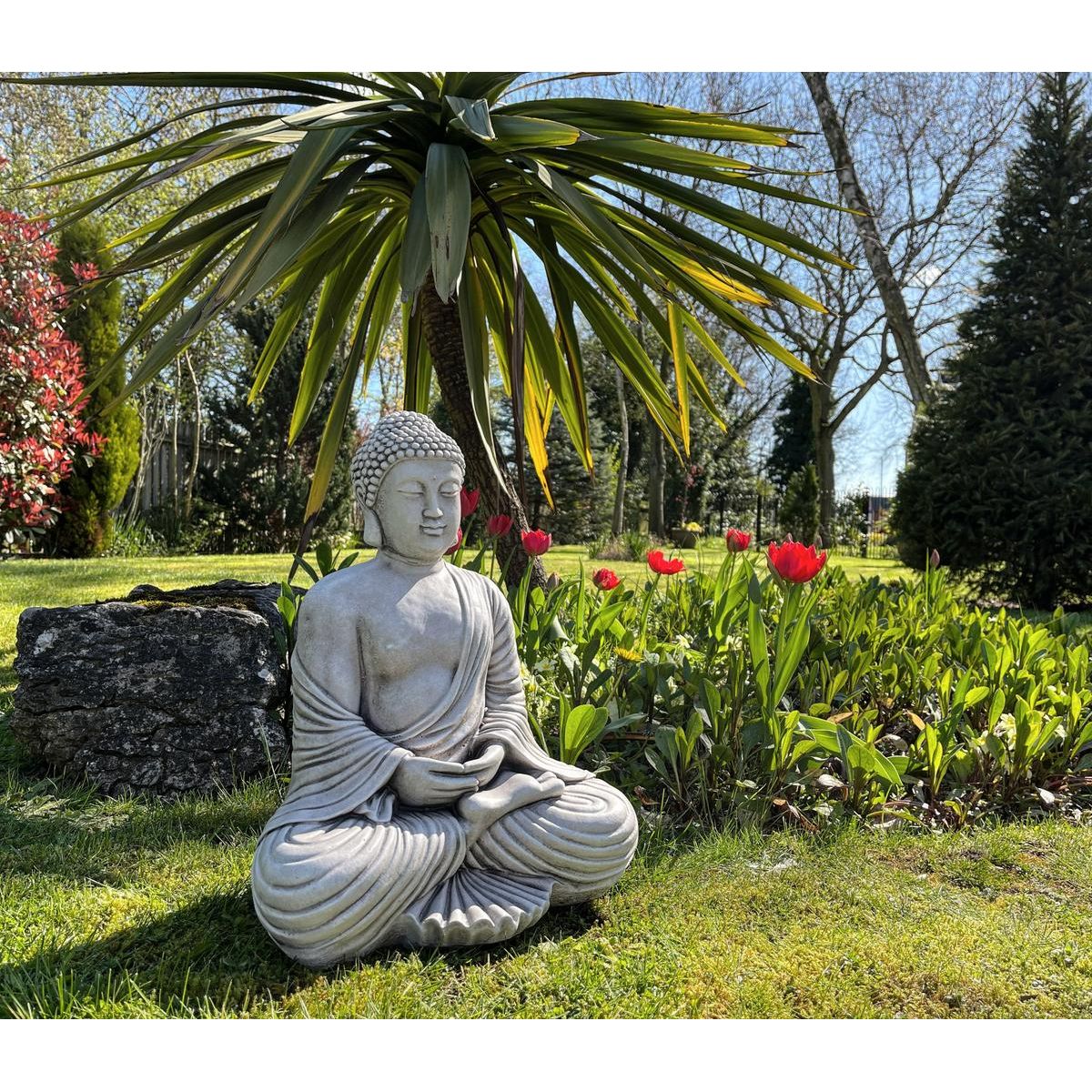 Stone Buddha statue in meditation pose, set on lush green grass with a tropical plant and red tulips in the background. Perfect for peaceful garden decor.